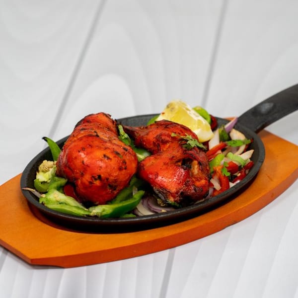 Two pieces of tandoori chicken with bell peppers, onions, and a lemon wedge in a cast iron pan.