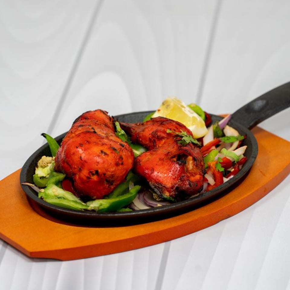 Two pieces of tandoori chicken with bell peppers, onions, and a lemon wedge in a cast iron pan.