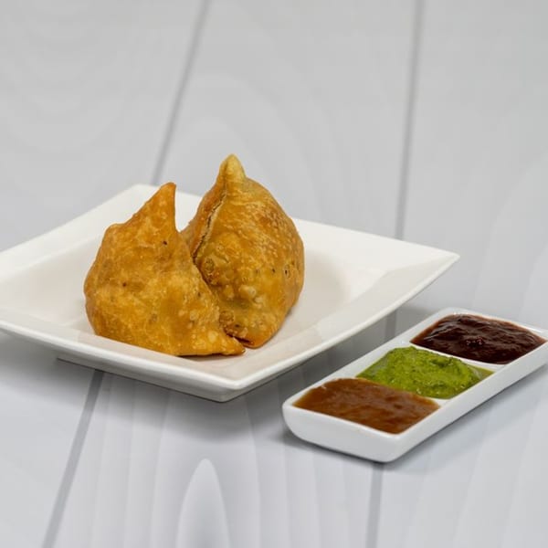 Two golden-brown samosas on a white plate, with a side of three colorful chutneys.