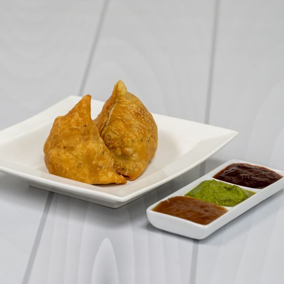 Two golden-brown samosas on a white plate, with a side of three colorful chutneys.